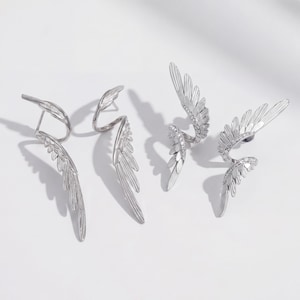 May include: Silver wing earrings with a detailed feather design. Some earrings are embellished with small, sparkling stones. The wings curve around the ear, creating a unique and elegant look. The earrings are displayed on a white surface.