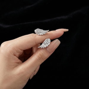 May include: A pair of silver-colored wing-shaped stud earrings. Each earring features a detailed feather design and is embellished with small, sparkling stones at the top. The earrings are displayed against a dark background.