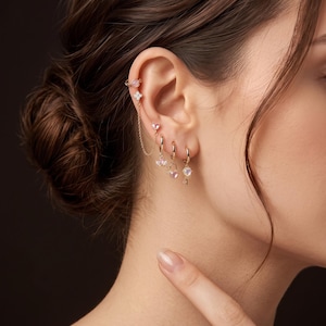 May include: A gold-toned ear cuff and earring set featuring a delicate chain connecting a top heart-shaped stud to a flower-shaped stud. Three small gold hoop earrings hang below, each adorned with a small, light pink heart-shaped charm.