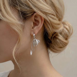 Op de afbeelding: Zilverkleurige oorhangers met een bloemmotief. De oorbellen hebben een gebogen, langwerpige vorm met een bloembladachtig element aan de bovenkant en vloeiende, lineaire details. De oorbellen worden op het oor gedragen en tonen hun elegante en moderne esthetiek.