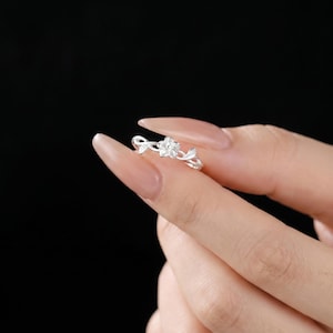 Könnte beinhalten: Ein silberner Ring mit einem zentralen, fünfblättrigen Blumendesign, das von zart geformten Blättern auf beiden Seiten der Ringschiene flankiert wird. Das Metall hat eine polierte, reflektierende Oberfläche und ein schmales Band.