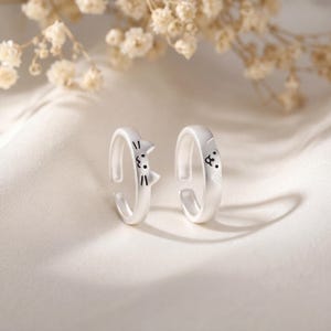 May include: Two silver-colored adjustable rings. One ring features a cat face design with black details, and the other ring has a dog face design. The rings are displayed on a white, textured surface with a blurred floral background.