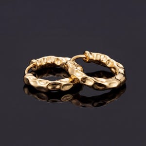 May include: A pair of gold-coloured hoop earrings with a textured, hammered design. The earrings are round and have a shiny finish, reflecting light. The background is dark, highlighting the jewellery.