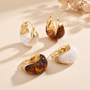 Puede incluir: Cuatro pendientes de aro con un diseño grueso y curvado con un marco de metal dorado pulido. Dos pendientes tienen una inserción de resina blanca veteada y dos tienen una inserción de resina marrón oscuro con patrón de carey.