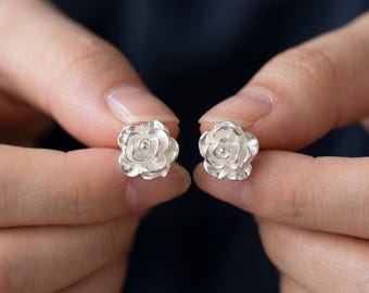 May include: A pair of silver rose-shaped stud earrings. The earrings are held between two hands against a dark background. The earrings are detailed with petals and a central bud, showcasing a floral design.