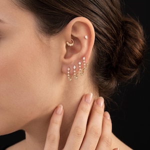 May include: A set of gold earrings featuring small hoops and dangling chains with clear stones. The jewellery includes a daith hoop, a flat stud, and three lobe earrings with marquise and round cut stones, creating a layered, delicate appearance.