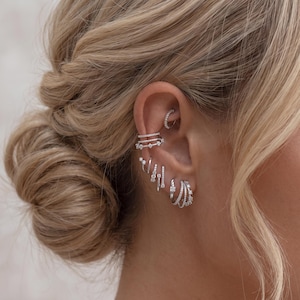 May include: A collection of silver-toned earrings and ear cuffs featuring clear stones. The set includes a small hoop in the rook, three stacked cuffs on the upper cartilage, two small dangling charms, and three layered hoops on the earlobe.