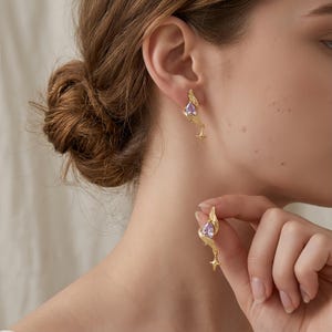 May include: A pair of gold-toned earrings featuring a teardrop-shaped light purple gemstone set within a curved, wing-like metal frame. Each earring terminates in a small, four-pointed star charm that hangs delicately from the base of the design.