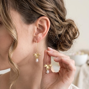 Peut inclure: Une paire de boucles d'oreilles pendantes dorées présentant un délicat motif de fleur à trois pétales en métal mat. Chaque boucle est suspendue à un fil fin et incurvé, terminée par une petite perle blanche en forme de goutte d'eau.