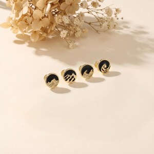 May include: Four gold-coloured stud earrings with black enamel designs of waves and mountains. The earrings are arranged on a cream-coloured surface, with dried flowers in the background. The earrings are small and delicate.