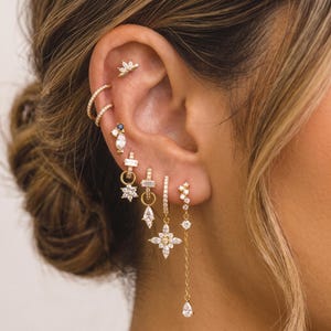 May include: A collection of gold-toned earrings featuring multiple piercings. The set includes a lotus-shaped stud, two thin pavé hoops, a small stud with a blue stone, and several dangling charms with clear crystals in star and teardrop shapes.