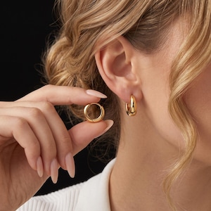 Op de afbeelding: Een paar kleine, gepolijste gouden oorringen met een glad, afgerond oppervlak. De ene oorring wordt tussen de vingers gehouden, terwijl de andere in de oorlel wordt gedragen. De ringen hebben een eenvoudig, doorlopend cirkelvormig ontwerp.