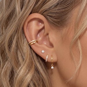 May include: A close-up of an ear adorned with gold earrings. The ear features a double-banded ear cuff, a small stud, a hoop earring with a dangling charm, and a small, straight earring. All earrings are gold and embellished with small, clear stones.