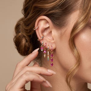 May include: A collection of gold-toned earrings worn in a single ear, featuring two upper ear cuffs with clear and multi-colored stones, and four dangling hoop earrings adorned with pink, clear, and multi-colored gemstones in various shapes.