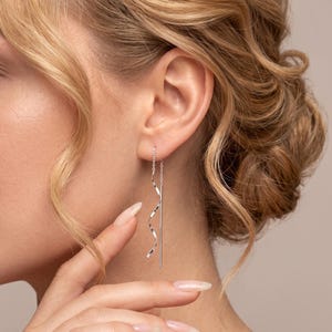 May include: A silver-toned threader earring featuring a delicate chain that transitions into a smooth, spiraling metal curve. The earring hangs vertically from the earlobe, showcasing a polished, reflective surface and a slender, straight post.