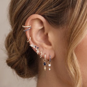 May include: A collection of gold-toned earrings featuring multiple small hoops and studs. The jewelry includes clear and blue stones in various shapes, such as marquise, pear, and round cuts, arranged along the ear cartilage and lobe.