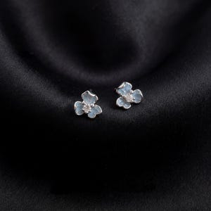 Puede incluir: Un par de pendientes de botón florales. Cada pendiente presenta un diseño de flor de tres pétalos azul claro con una pequeña gema redonda y transparente en el centro. Los pendientes están colocados sobre un fondo de satén negro.