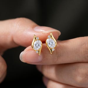 May include: A pair of gold stud earrings featuring a central oval-cut clear gemstone flanked by two smaller round-cut clear gemstones, set within a curved, leaf-like gold frame that tapers to points at the top and bottom of each earring.