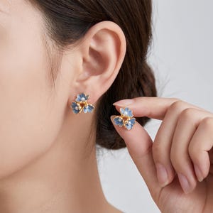 Könnte beinhalten: Ein Paar goldfarbene Ohrstecker in Form von dreiblättrigen Blumen mit hellblauen Emaille-Blütenblättern und einer kleinen weißen Perle in der Mitte jeder Blume. Die Ohrringe haben ein metallisches Gold-Finish und ein filigranes Blumendesign.