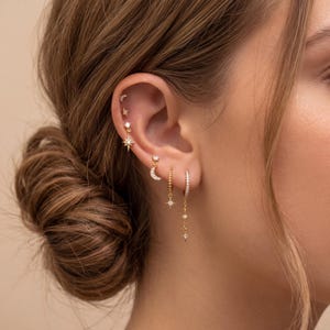 Puede incluir: Una colección de pendientes dorados, que incluyen un pendiente de estrella, un pendiente de media luna y un pendiente de aro con estrellas colgantes. Los pendientes se llevan en la oreja de una persona con cabello castaño. Los pendientes están adornados con pequeñas piedras brillantes.