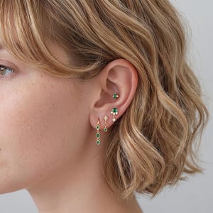 May include: A set of four gold-toned earrings featuring green gemstones. The collection includes a small rectangular stud, a round stud with accent stones, a diamond-shaped drop earring, and a dangling hoop earring with three green stones.