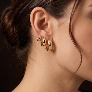 May include: Three gold hoop earrings of varying sizes arranged in a row on an earlobe. The largest hoop features small, engraved star motifs with tiny embedded stones, while the two smaller hoops have a smooth, polished gold finish.