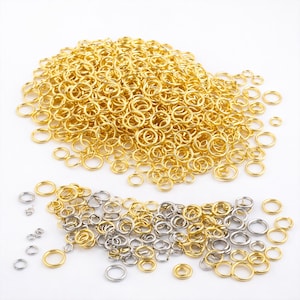 Könnte beinhalten: Eine Ansammlung gold- und silberfarbener Biegeringe, die auf einer weißen Oberfläche verstreut sind. Die Biegeringe sind in verschiedenen Größen erhältlich und werden für die Schmuckherstellung und zum Basteln verwendet. Die goldenen Ringe sind in einem großen Haufen angeordnet.