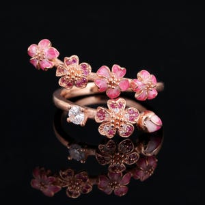 Może przedstawiać: Pierścionek z różowego złota z kwiatowym wzorem. Pierścionek posiada różowe emaliowane kwiaty z czerwonymi kamieniami szlachetnymi w środku i przezroczysty akcent z kamieni szlachetnych. Pierścionek jest na czarnym tle.