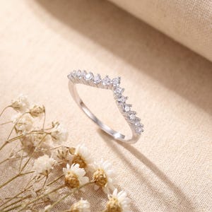 May include: A silver-colored ring with a V-shaped design, adorned with small, clear gemstones. The ring is set against a neutral background with dried flowers, highlighting its delicate and elegant appearance. A piece of jewelry.