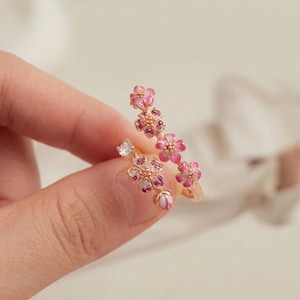K&ouml;nnte beinhalten: Ein ros&eacute;goldener Ring mit floralem Design, rosa Emaillebl&uuml;ten und kleinen, funkelnden Edelsteinen. Der Ring hat eine zarte, feminine &Auml;sthetik, mit einer Blumengruppe, die sich um den Finger windet.