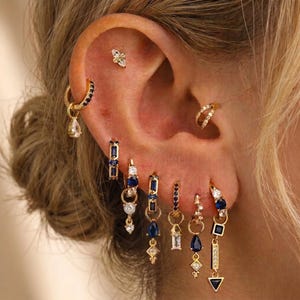 Könnte beinhalten: Eine Sammlung goldfarbener Ohrringe in mehreren Ohrlöchern, bestehend aus verschiedenen Designs wie kleinen Creolen, hängenden Anhängern und Steckern. Der Schmuck ist mit tiefblauen Edelsteinen und klaren weißen Kristallen in verschiedenen Formen besetzt.
