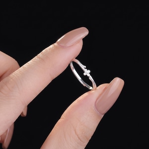 May include: A thin silver-toned ring featuring a delicate band that mimics the appearance of a small twig or branch. The center of the ring is adorned with a tiny, four-petaled flower motif, which is set with small, clear, sparkling stones.