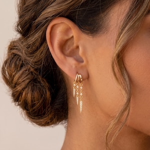May include: Three gold hoop earrings worn in a single earlobe. Each hoop features a dangling chain element adorned with small, clear, round-cut stones. The central and longest chain ends in a pointed, stone-encrusted charm, while the others are shorter.