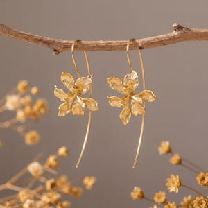 Peut inclure: Une paire de boucles d'oreilles pendantes dorées. Chaque boucle d'oreille présente un motif détaillé en forme de fleur avec de longs fils incurvés. Les boucles d'oreilles sont suspendues à une fine branche brune sur un fond neutre, avec des fleurs séchées au premier plan.