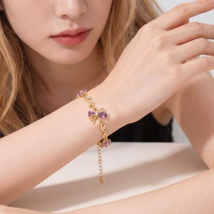 Op de afbeelding: Goudkleurige armband met een strik- en bloemmotief met paarse edelstenen. De armband heeft een fijne ketting en is versierd met kleine, heldere kristallen. De sieraden worden om de pols gedragen.