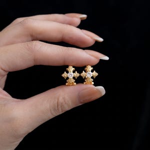 Può includere: Un paio di orecchini a cerchio dorati con un design decorativo a forma di croce sulla parte anteriore. Ogni croce è impreziosita da una singola pietra preziosa trasparente a taglio rotondo al centro, su una superficie in metallo dorato.