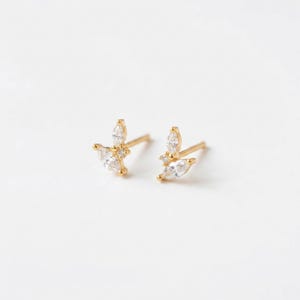 Può includere: Un paio di orecchini a lobo color oro. Ogni orecchino presenta un gruppo di pietre di zirconia cubica trasparenti, taglio marquise e rotondo, disposte in un delicato design a forma di foglia. Gli orecchini sono su uno sfondo bianco.
