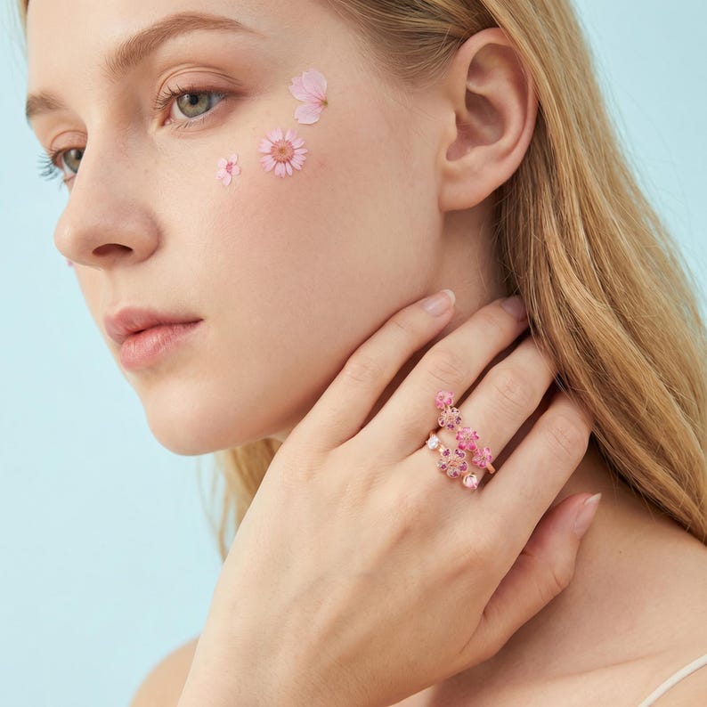K&ouml;nnte beinhalten: Ein ros&eacute;goldener Ring mit rosa floralen Designs und kleinen Edelsteinen. Der Ring zeigt eine Ansammlung von Blumen und wird an einem Finger getragen. Rosa Bl&uuml;tenbl&auml;tter sind auf der Wange platziert.