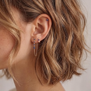 May include: An ear with three gold earrings. One is a small stud, another a hoop with a small white gem, and the third a chain earring with blue rectangular gems. A small blue stud is in the upper cartilage of the ear.