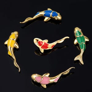 May include: Five gold-toned metal koi fish pins, each featuring colorful enamel inlays of blue, yellow, red, green, and pink. Each fish has a small, clear stone set near the head, with detailed fins and tails, arranged in a circular formation.