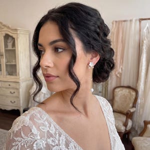 以下が含まれることがあります： 白い花のイヤリングをつけた女性。イヤリングには揺れるパールとゴールドチェーンが付いています。背景には、白いキャビネットとレースのドレスがあるヴィンテージスタイルの部屋が写っています。