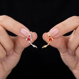 Puede incluir: Dos pequeños colgantes de peces koi, uno plateado y otro dorado, cada uno con detalles rojos y blancos o rojos y dorados. Los colgantes se sostienen sobre un fondo negro.