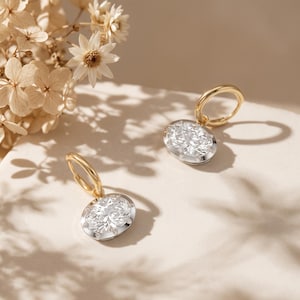 May include: A pair of hoop earrings featuring oval-shaped clear stones set in silver-toned metal, suspended from polished gold-toned hoops. The earrings are displayed with a cluster of dried white flowers, creating a bright and clean aesthetic.