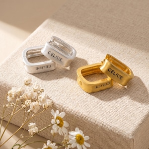 May include: Silver and gold-coloured square earrings with the word "CELIOE" engraved. The earrings are displayed on a beige surface with white flowers in the foreground. The earrings have a modern, minimalist design, suitable for everyday wear.