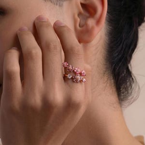 K&ouml;nnte beinhalten: Ein ros&eacute;goldener Ring mit floralem Design. Der Ring hat zwei B&auml;nder, die jeweils mit rosa bl&uuml;tenf&ouml;rmigen Edelsteinen verziert sind. Die Bl&uuml;ten haben verschiedene Rosat&ouml;ne und sind gruppiert, was eine zarte und feminine &Auml;sthetik erzeugt.