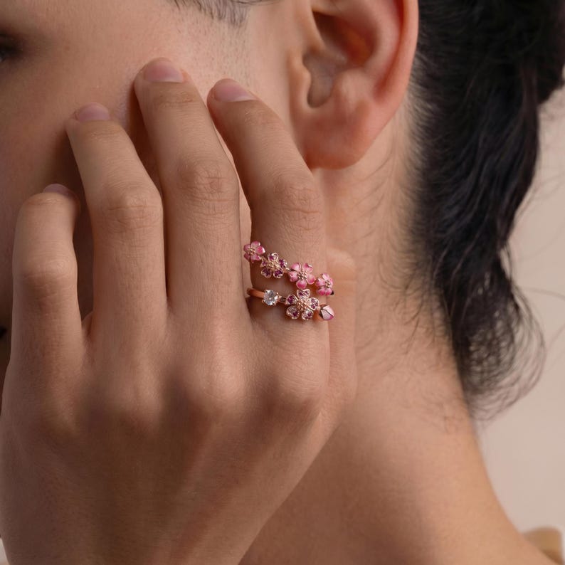 K&ouml;nnte beinhalten: Ein ros&eacute;goldener Ring mit floralem Design. Der Ring hat zwei B&auml;nder, die jeweils mit rosa bl&uuml;tenf&ouml;rmigen Edelsteinen verziert sind. Die Bl&uuml;ten haben verschiedene Rosat&ouml;ne und sind gruppiert, was eine zarte und feminine &Auml;sthetik erzeugt.