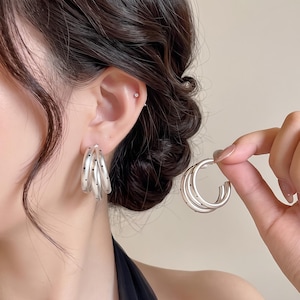 May include: A pair of silver-toned hoop earrings featuring a triple-layered design. Each hoop is crafted with a smooth, matte finish and accented with small, evenly spaced black dots along the outer surface of the metal bands.