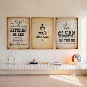 Op de afbeelding: Drie ingelijste wandkunstwerken met keukenthema. Het linkerframe leest "KITCHEN RULES", het middelste frame zegt "cook with love" en het rechterframe zegt "CLEAN AS YOU GO". De frames zijn bruin met een vintage papieren achtergrond.