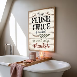 Op de afbeelding: Een houten bord met de tekst "Please FLUSH TWICE if needed, we won't judge thanks!" wordt in een badkamer getoond. Het bord heeft een rustiek ontwerp met katoen- en bladillustraties. Een badkuip dienblad bevat een kaars, een pot en een klein schaaltje.