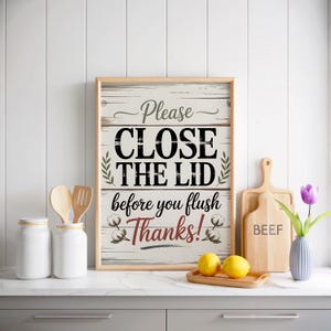 Puede incluir: Se muestra un letrero enmarcado con el texto "Please CLOSE THE LID before you flush Thanks!". El letrero está sobre un fondo de madera blanca. También se ven dos frascos blancos, utensilios de madera, limones y una tabla de cortar.